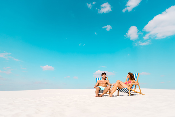 sonriente joven pareja sentada en tumbonas en la playa de arena
 - Foto, imagen