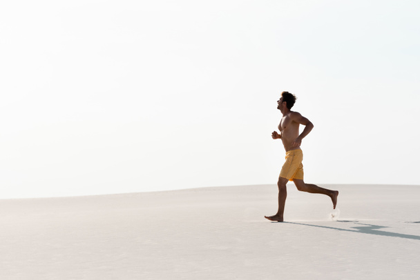 άνδρας με μαγιό σορτς και μυϊκό κορμό τρέχει σε αμμώδη παραλία - Φωτογραφία, εικόνα