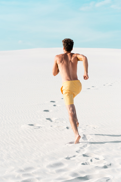 θέα προς τα πίσω του άνδρα με μαγιό σορτς με μυϊκό κορμό που τρέχει στην αμμώδη παραλία - Φωτογραφία, εικόνα