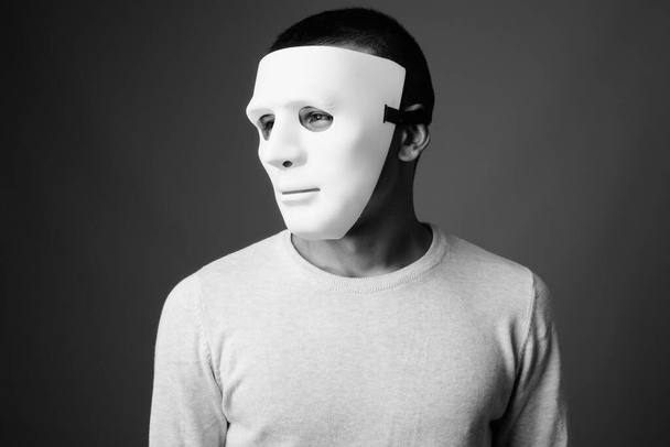Studio shot van jonge man met witte masker tegen grijze achtergrond in zwart-wit - Foto, afbeelding