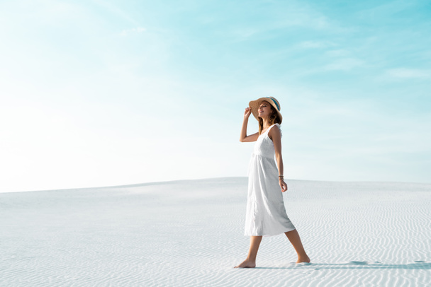zijaanzicht van lachend mooi meisje in witte jurk en strohoed wandelen op zandstrand met blauwe lucht - Foto, afbeelding