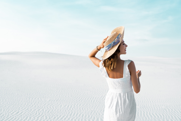 back view of beautiful girl in white dress and straw hat on sandy beach with blue sky - Zdjęcie, obraz