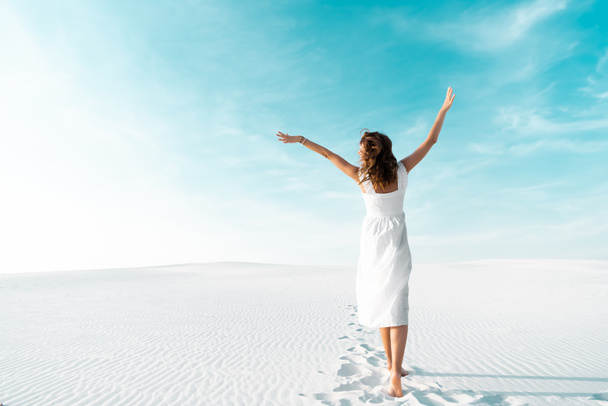 back view of beautiful girl in white dress with hands in air on sandy beach with blue sky - Foto, afbeelding