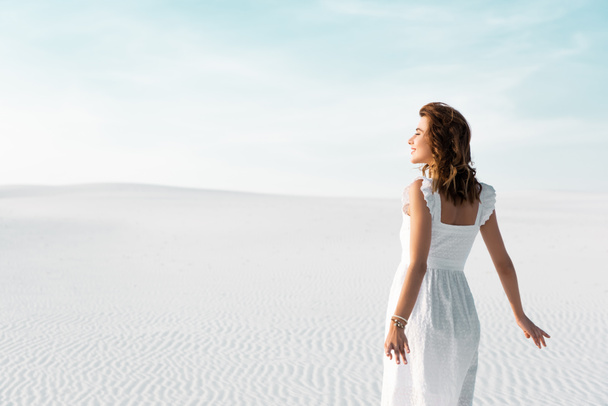 back view of smiling beautiful girl in white dress on sandy beach with blue sky - Zdjęcie, obraz