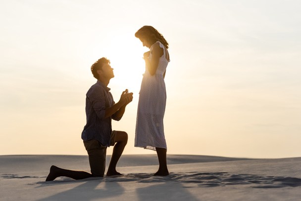 oldalnézetben fiatal férfi csinál házassági ajánlat barátnője homokos tengerparton naplementekor - Fotó, kép