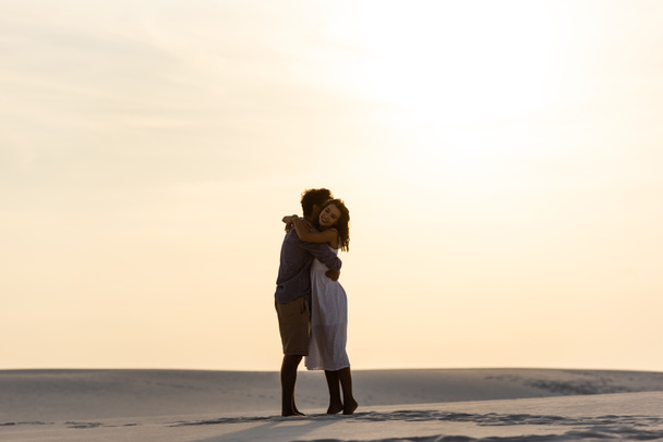 πλαϊνή άποψη του νεαρού ζευγαριού αγκαλιάζει στην αμμώδη παραλία στο ηλιοβασίλεμα - Φωτογραφία, εικόνα