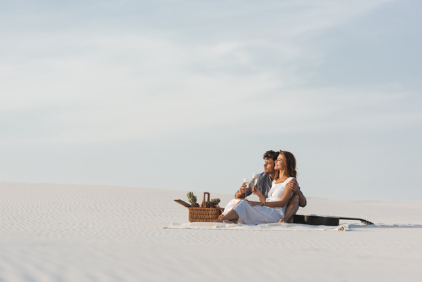 young couple drinking champagne while sitting on blanket with basket of fruits and acoustic guitar on beach - Фото, зображення