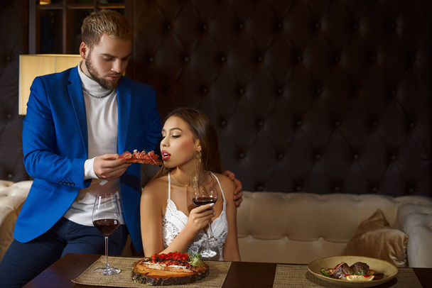 restaurant and holiday concept - Luxurious couple eating at restaurant.The girl eats from the hands of a guy bruschetta. - Foto, afbeelding