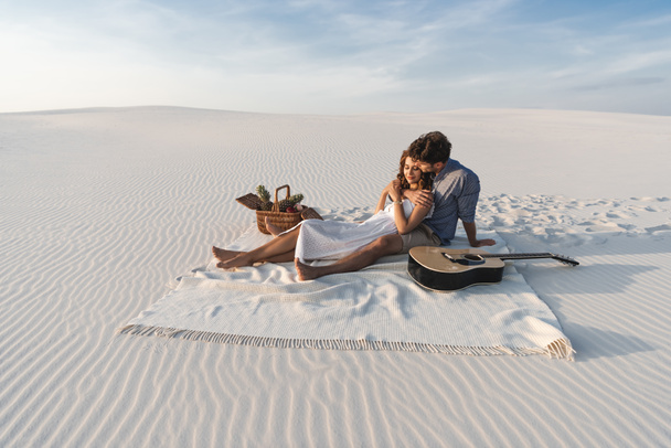 young couple hugging while sitting on blanket with basket of fruits and acoustic guitar on beach - Foto, Imagen