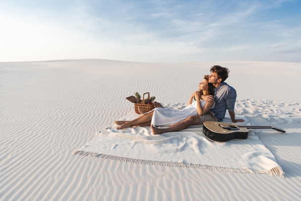 young couple hugging while sitting on blanket with basket of fruits and acoustic guitar on beach - Фото, зображення
