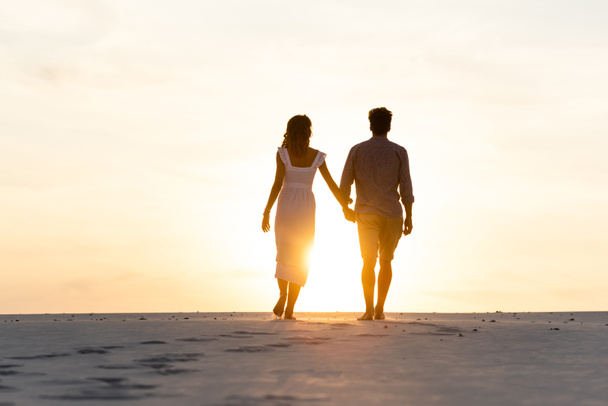 silhouettes of man and woman holding hands while walking on beach against sun during sunset - Foto, imagen