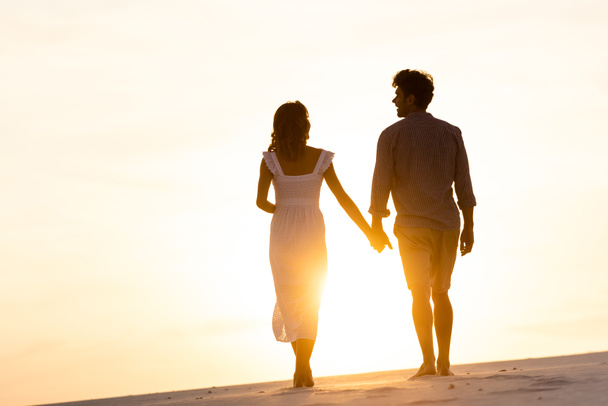 silhouettes of man and woman holding hands while walking on beach against sun during sunset - Photo, Image