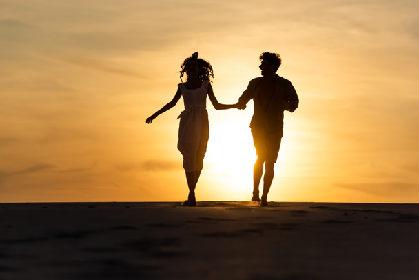 силуэты мужчины и женщины, бегущие по пляжу от солнца на закате
 - Фото, изображение