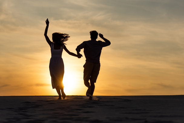 silhouettes of man and woman running on beach against sun during sunset - Photo, Image