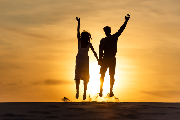 silhouettes of man and woman jumping on beach against sun during sunset - 写真・画像