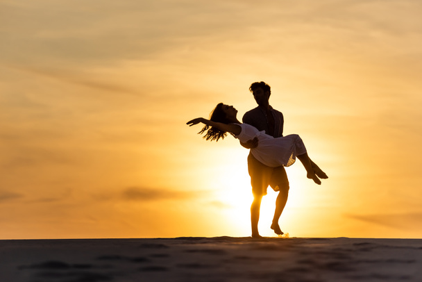 silhouettes of man spinning around woman on beach against sun during sunset - Photo, Image