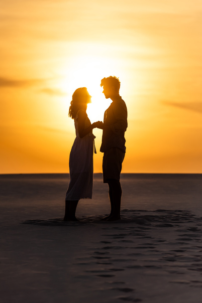 side view of silhouettes of man and woman holding hands on beach against sun during sunset - Photo, Image