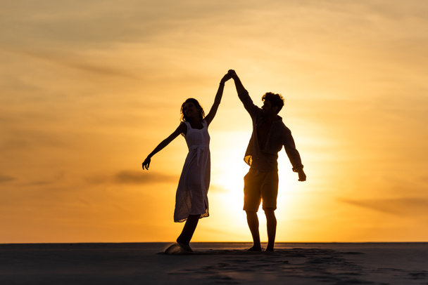silhouettes of man and woman dancing on beach against sun during sunset - Photo, Image