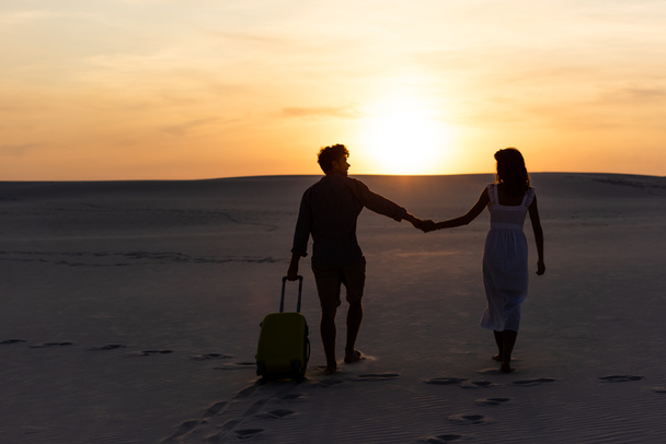 back view of couple walking on beach while holding hands with travel bag at sunset - Foto, imagen