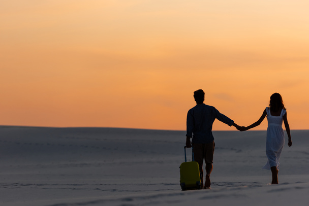 vue arrière du couple marchant sur la plage tout en se tenant la main avec un sac de voyage au coucher du soleil
 - Photo, image