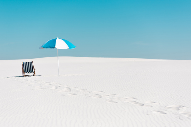 kansituoli ja sateenvarjo hiekkarannalla jälkiä vastaan kirkas sininen taivas
 - Valokuva, kuva