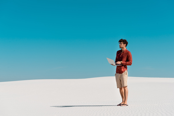 young freelancer on sandy beach with laptop against clear blue sky - Foto, Bild