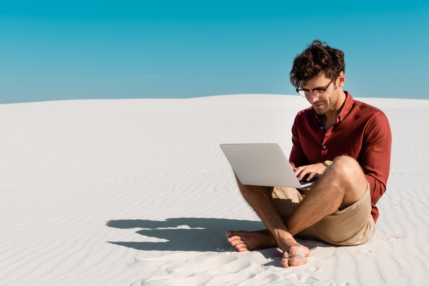 young freelancer on sandy beach using laptop against clear blue sky - Фото, изображение