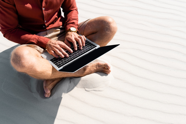 bijgesneden weergave van jonge freelancer zitten op zandstrand met laptop - Foto, afbeelding