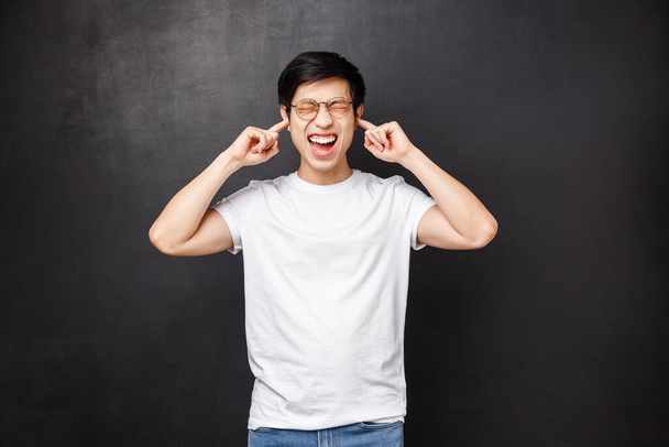 Lifestyle and people concept. Bothered and anguished young asian guy screaming from discomfort and distress as hearing loud noise, close ears with fingers shut eyes disturbed, stand black background - Photo, Image