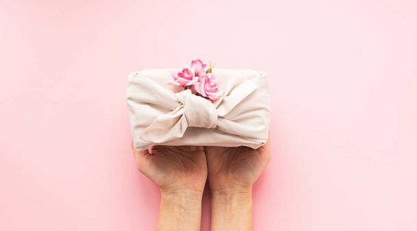 Female hands holding a holiday gift packed fabric in the manner of Furoshiki on a fashionable pink background. - Photo, Image
