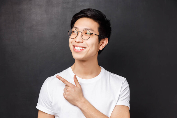 Nahaufnahme Porträt eines verträumten netten asiatischen Typen mit Brille, der etwas Cooles sieht, mit zufriedenem Gesichtsausdruck und strahlendem Lächeln in die linke obere Ecke zeigt und schaut, schwarzer Hintergrund - Foto, Bild