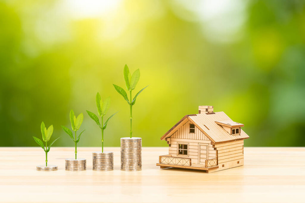 Монета стоїть і модель будинку зі зростаючим листям на дерев'яному столі на фоні зеленого дерева, концепція іпотеки
 - Фото, зображення