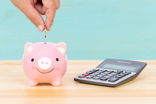 Varkensbank met hand inzet munt en rekenmachine, op bureau, over lichtblauwe houten achtergrond, geldbesparend concept - Foto, afbeelding