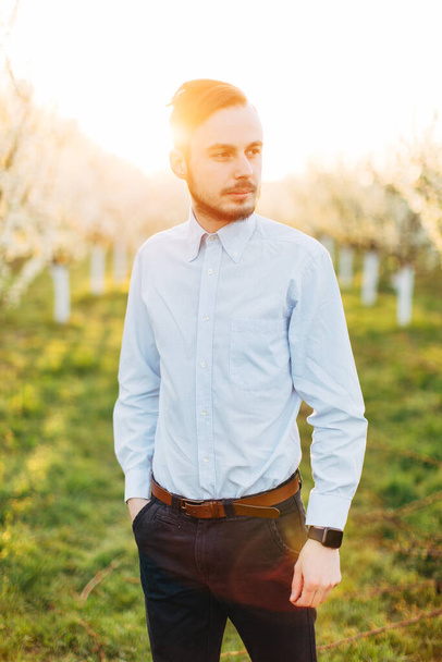 Outdoors portrait of bearded stylish groom man in light shirt and dark pants. Handsome smiling man outdoors, walking in spring garden with blooming trees, sunny day. - 写真・画像