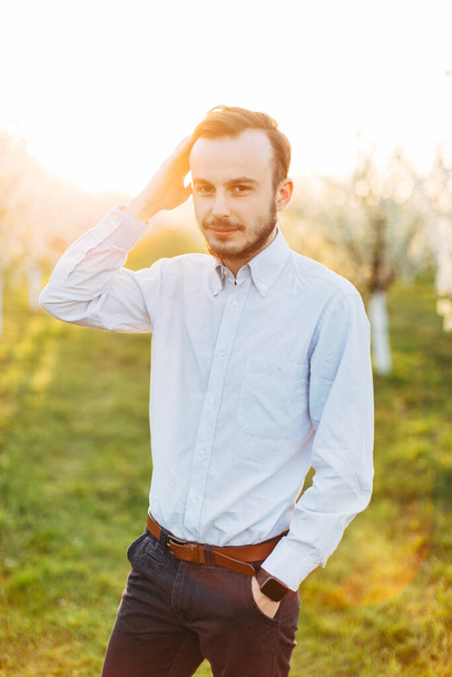 Young man weared in smart casual suit on background of the blooming garden and flowering trees. Fashion style guy happy groom in white shirt posing over the garden and sunrise background - Photo, Image