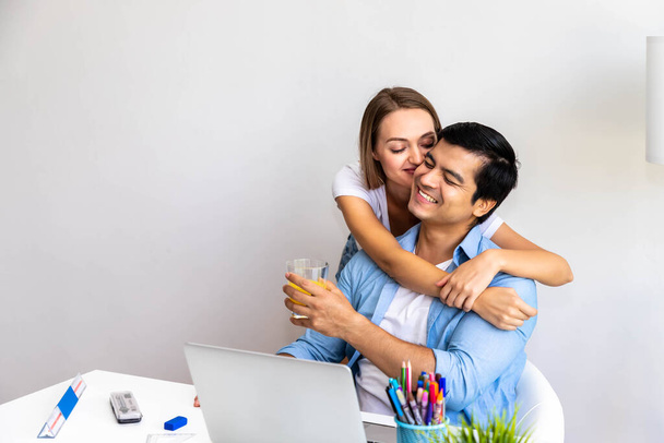 Чоловік працює вдома за допомогою комп'ютерного ноутбука, в той час як жінка дружина подає апельсиновий сік, обіймаючи і цілуючи його
 - Фото, зображення