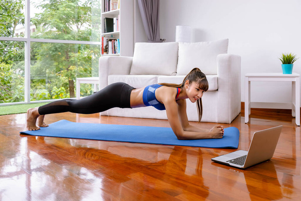 Atractiva mujer asiática en ropa deportiva practicando yoga y viendo lección de tutorial en computadora portátil haciendo ejercicio de tablón en la sala de estar en casa
 - Foto, imagen