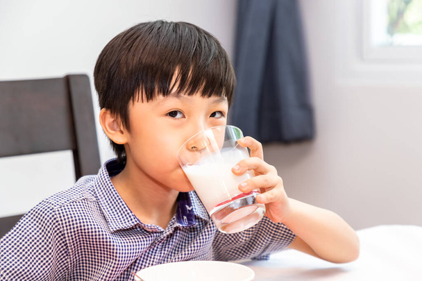Adorável jovem asiático feliz tomando café da manhã e bebendo leite em casa pela manhã; olhando para a câmera
 - Foto, Imagem