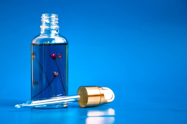 Tropferglasflasche mit Blumen in Mock-Up. Kosmetische Pipette auf blauem Hintergrund. Nagelhaut-Öl, Anti-Aging-Serum in Glasflasche mit Tropfer Hautpflegeessenz für schöne gesunde Haut - Foto, Bild