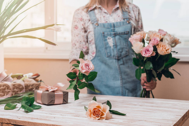 Χέρια της νεαρής γυναίκας ανθοπωλείο που εργάζονται με φρέσκα λουλούδια κάνοντας μπουκέτο από ροζ τριαντάφυλλα στο τραπέζι. Κλείσε.. - Φωτογραφία, εικόνα