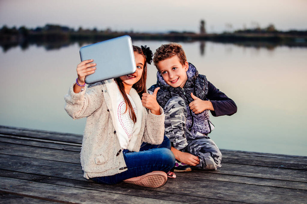 Két fiatal aranyos kis barátok, fiú és lány szórakozik, és szelfit csinál a blogolás digitális táblagépen, miközben ül a tó este. A fiú és a lány ujjal mutogatnak. Barátság - Fotó, kép