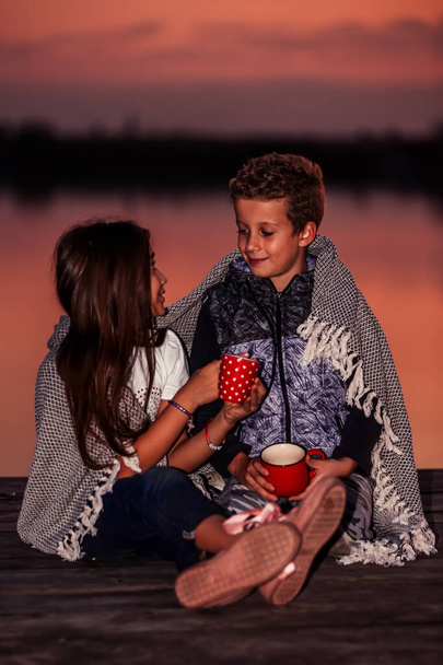 Dos jóvenes amiguitos lindos, niño y niña hablando mientras están sentados cubiertos con manta junto al lago durante el atardecer. Enfoque selectivo
 - Foto, imagen