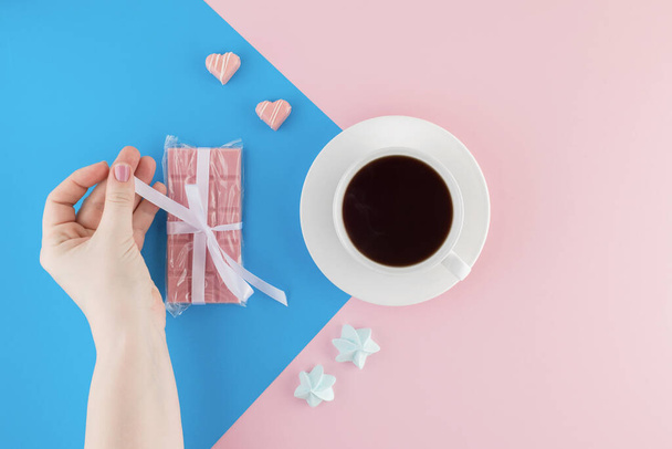 Female hand holds a cup of freshly brewed coffee on beautiful pink and bright blue pastel background. Fresh desserts. Meringue. Pink Chocolate and heart shaped candies. Top view. Flat lay.  - Фото, изображение