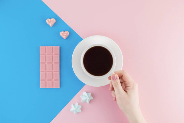 Female hand holds a cup of freshly brewed coffee on beautiful pink and bright blue pastel background. Fresh desserts. Meringue. Pink Chocolate and heart shaped candies. Top view. Flat lay.  - Foto, imagen