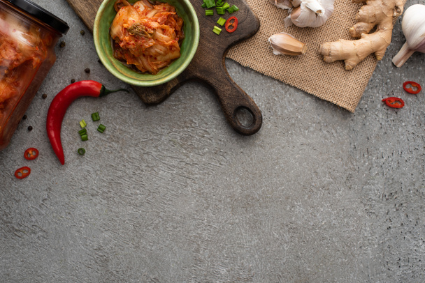 top view of kimchi in bowl and jar near chili pepper, garlic and ginger on concrete surface - Foto, afbeelding