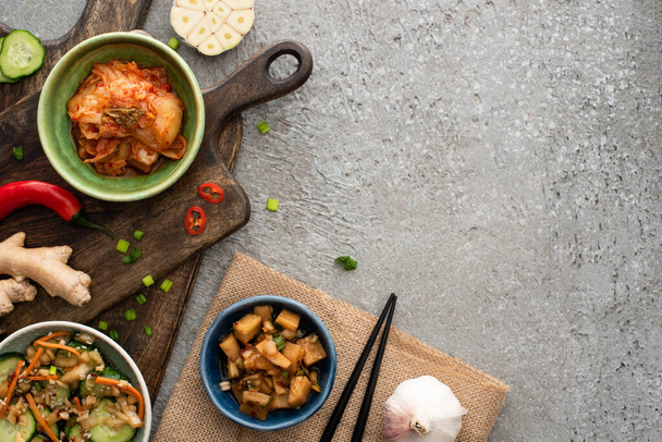 bovenaanzicht van kommen met kimchi op snijplanken bij eetstokjes, knoflook, gember en chili peper op betonnen ondergrond - Foto, afbeelding