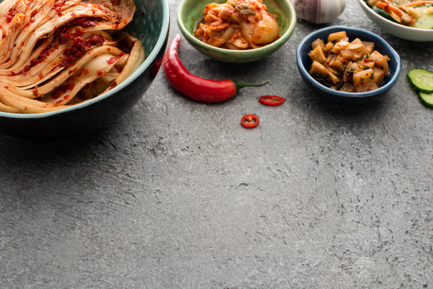 bowls with tasty kimchi near chili pepper and garlic on concrete surface - Фото, зображення
