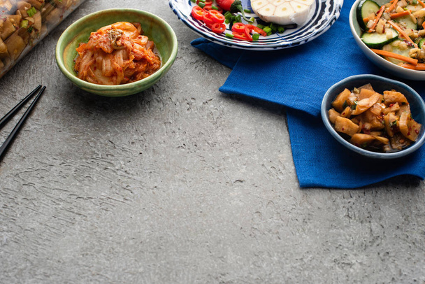 kimchi sabroso en cuencos y frasco en tela azul cerca de palillos en la superficie de hormigón
  - Foto, Imagen