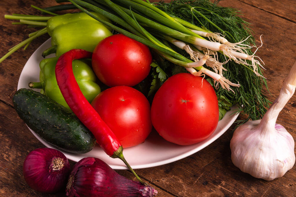 Verduras frescas, tomates, tomates cherry, pimientos verdes, chiles, cebollas, pepinos, ajo y hierbas en un plato en una mesa rústica de madera - estilo rural, concepto de comida saludable y orgánica
 - Foto, Imagen