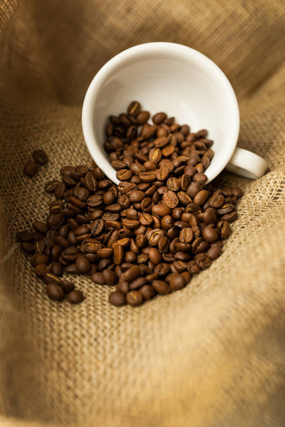 A white cup with scattered coffee beans on burlap. A scattering of coffee beans with a cup. Coarse fabric. - Fotó, kép